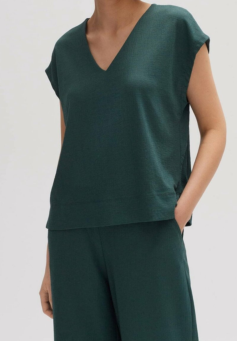 Top vert foncé à col en V, à manches courtes, en tissu texturé, avec une coupe décontractée et des fentes latérales pour plus de liberté de mouvement.