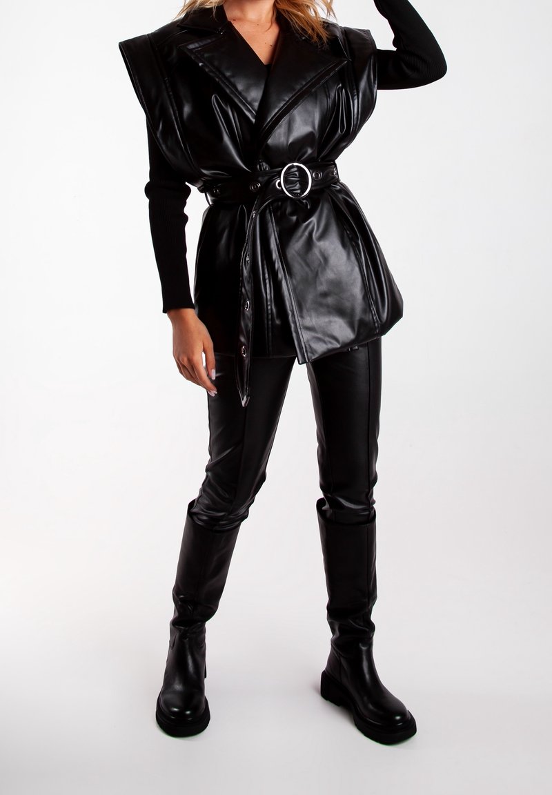 Femme portant un gilet en cuir noir sans manches surdimensionné avec ceinture, un haut noir à manches longues, un pantalon en cuir et des bottes noires montant jusqu'aux genoux sur fond blanc.