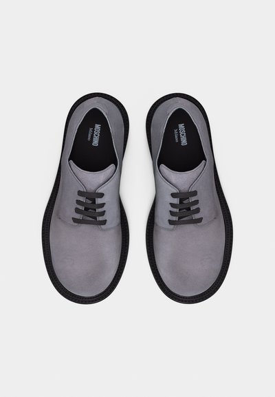 Paire de chaussures Oxford en daim gris avec lacets noirs et semelles noires, vue de dessus, marque "Moschino Milano" à l'intérieur.