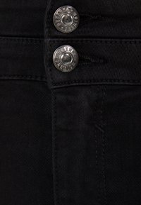 Jeans de mezclilla negra con un primer plano de dos botones de metal plateado y costuras visibles, que resaltan la textura de la tela y los detalles del diseño.