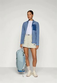 Mulher vestindo uma jaqueta azul, camisa branca, calções bege e botas de caminhada, segurando uma mochila azul clara contra um fundo liso.