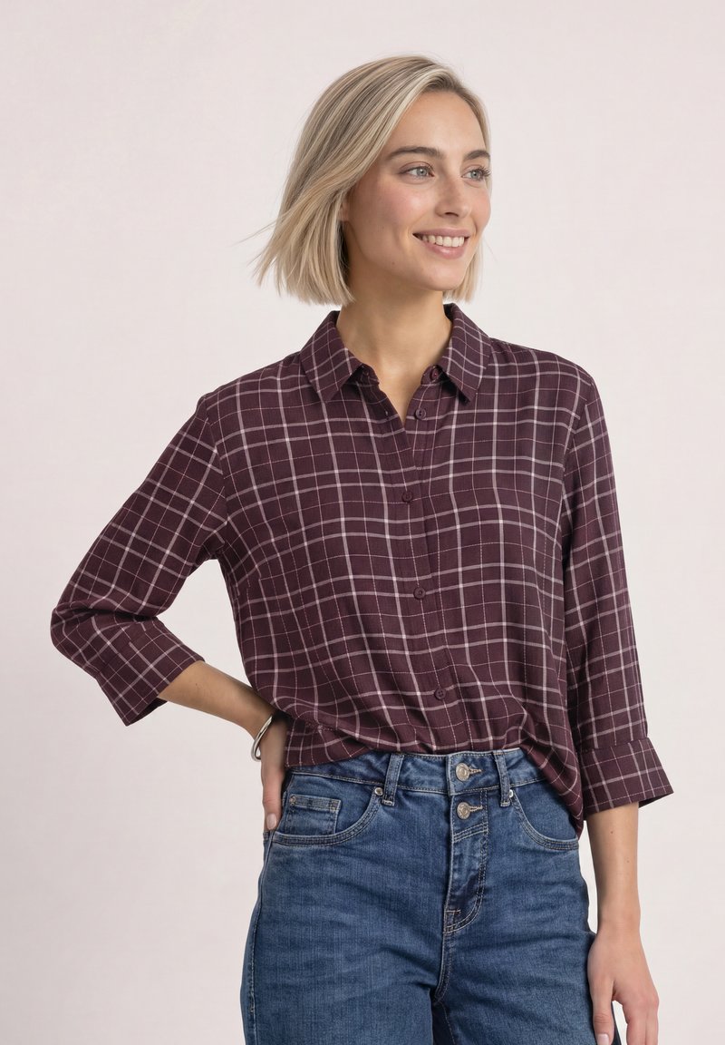 Femme souriante portant une chemise à carreaux violette avec les manches retroussées et un jean bleu taille haute, debout avec une main sur la hanche.