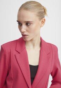 Taillierter Blazer aus leuchtend pinkem Stoff mit glatter Textur, Ein-Knopf-Verschluss und scharfen Revers, getragen über einem schwarzen Top.
