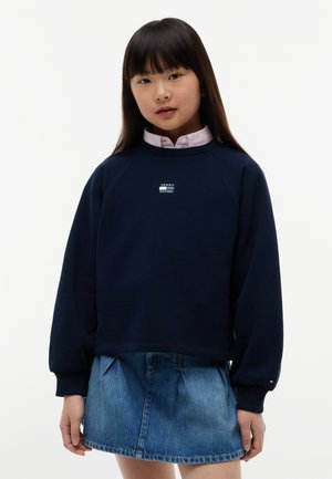 Young girl with long dark hair wearing a navy sweatshirt over a pink collared shirt and a denim skirt, standing against a plain white background.