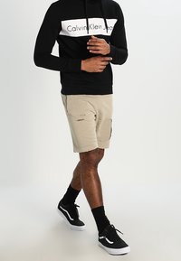 Felpa nera con logo bianco "Calvin Klein Jeans", pantaloncini beige strappati con toppe, sneakers nere e calzini neri.
