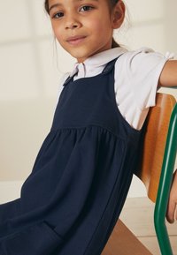 Granatowa sukienka z miękkiego materiału, z regulowanymi ramiączkami, marszczonym talią i kieszeniami. Modelka siedzi na drewnianym krześle.