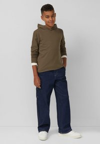 Brauner Kapuzenpullover, dunkelblaue Jeanshose, weiße Sneaker. Der Pullover hat lange Ärmel mit Bündchen und eine lässige, lockere Passform.