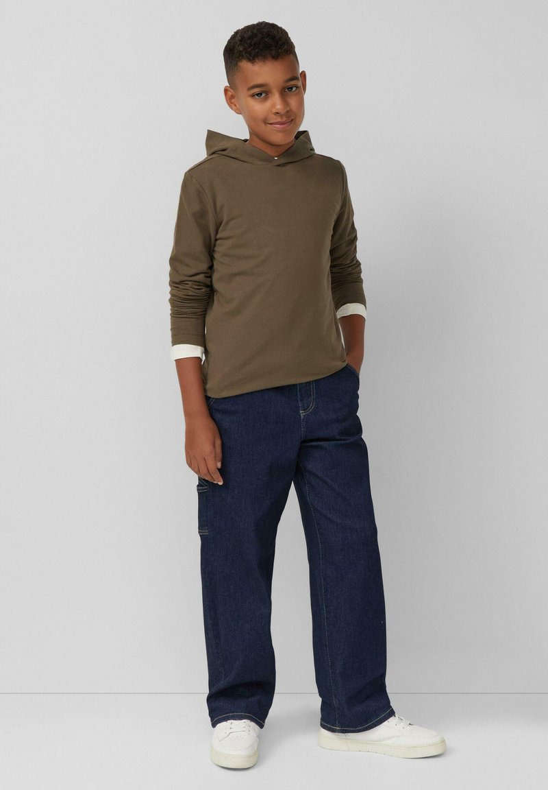 Brauner Kapuzenpullover, dunkelblaue Jeanshose, weiße Sneaker. Der Pullover hat lange Ärmel mit Bündchen und eine lässige, lockere Passform.