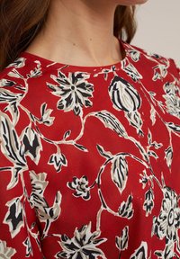 Tessuto rosso con un motivo floreale in nero e crema, con collo rotondo e texture liscia. Nessun sistema di chiusura visibile o dettagli decorativi.