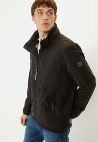 Veste légère noire avec un col haut, plusieurs poches zippées et un tirette de fermeture éclair orange contrastante. Détails en tissu texturé.