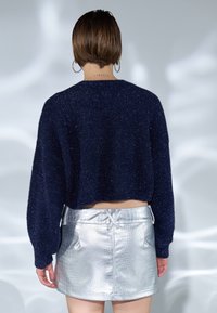 Suéter recortado de color marino con una textura brillante, combinado con una mini falda plateada y brillante con un estampado de cocodrilo. Presenta bolsillos laterales y un cordón ajustable.