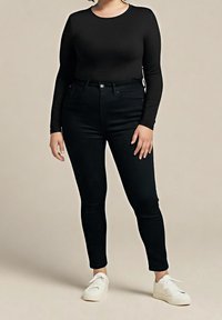 Femme portant un haut noir à manches longues, un jean skinny taille haute noir et des baskets blanches, debout devant un fond uni.