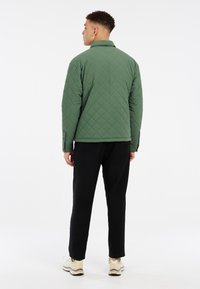 Gequilted groene jas met een kraag, met ruitenpatronen. Gepaard met zwarte broeken en lichte sneakers, gezien vanuit de achterkant.