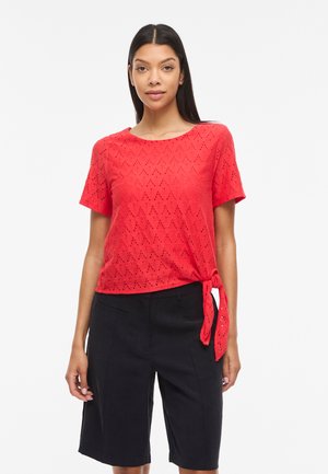 Γυναίκα που φοράει κόκκινο κοντομάνικο τοπ με τρύπες, δεμένο στο πλάι, και μαύρες, ραμμένες μέχρι το γόνατο σορτς, στέκεται μπροστά σε απλό φόντο.