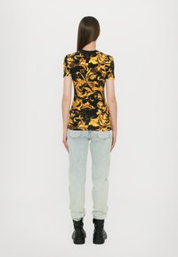 Kortermet svart topp med gul ornamentert mønster, kombinert med lyseblå denimjeans og svarte ankelstøvler. Vist bakfra.