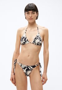 Rövid sötét hajú nő fekete-fehér virágmintás, oldalt megkötős, pánt nélküli bikiniben áll egy egyszerű, világos háttér előtt.