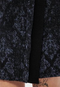 Gros plan sur une jupe en tissu foncé texturé avec une fente à l'avant, laissant apparaître un tatouage de visage sur la cuisse en dessous.