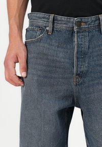 Vaqueros de mezclilla azul oscuro con corte recto, que cuentan con una cremallera de botones, costuras naranjas y un diseño de cinco bolsillos. La textura parece resistente y clásica.