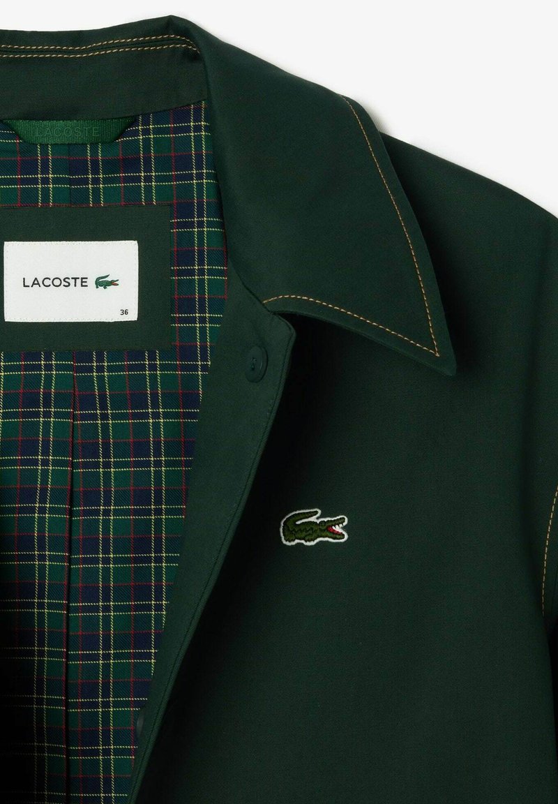 Veste Lacoste vert foncé avec un grand col, dotée d'un intérieur à carreaux rouge, jaune et vert. Comprend un logo crocodile brodé.
