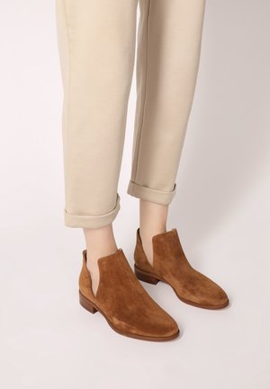 Beige rolled-up pants worn with brown suede ankle boots featuring cut-out sides and low wooden heels on a light background.