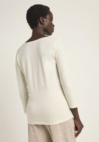 Helles, cremefarbenes Langarmoberteil mit weicher Textur, Rundrücken und figurbetonter Silhouette, kombiniert mit gestreiften braunen und weißen Hosen.