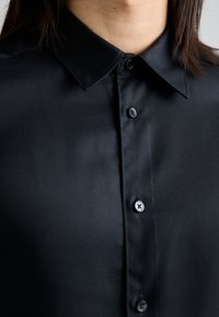 Camicia nera con bottoni, realizzata in tessuto morbido, caratterizzata da un colletto a punta e tre bottoni abbinati lungo la patta frontale, con una leggera lucentezza.