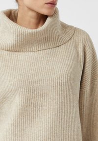 Beżowy sweter z prą�żkowanego dzianiny z wysokim kołnierzem, charakteryzujący się miękką fakturą i luźnym krojem. Zauważalny poziomy detal prążkowania.