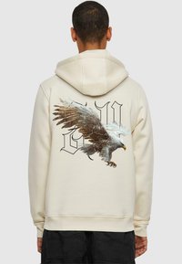 Cremfarbener Hoodie mit einem großen Adlerdesign und stilisierten Buchstaben auf dem Rücken. Weicher Stoff, lockere Passform und verstellbare Kapuze.