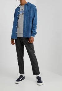 Hombre vistiendo una camisa de botón a rayas azules sobre una sudadera gris con gráfico, pantalones a rayas negros y zapatillas azul marino con suelas blancas.