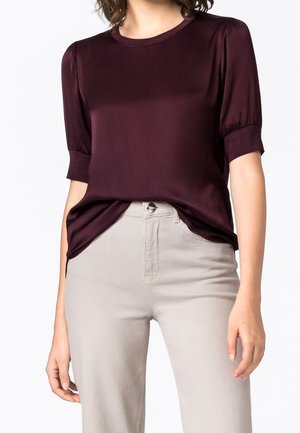 Vrouw draagt een donkerpaarse korte mouwen blouse, ingestopt in lichtbeige, hooggesneden broek met knoopsluiting.