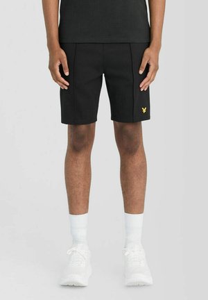 Mandlig model iført sorte shorts med et lille gult logo på venstre ben, kombineret med hvide sokker og hvide sneakers, stående foran en ensfarvet baggrund.