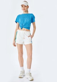 Blå kortærmet top med knude foran, parret med hvide shorts med sideløsninger i blå og grøn. Tilbehøret inkluderer en hvid skyggehue.