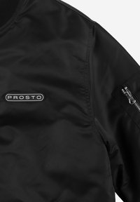Czarna satynowa kurtka bomberka z prążkowanym kołnierzem, wyraźnym logo "PROSTO" oraz zamkiem błyskawicznym na lewym rękawie. Gładka tekstura, minimalistyczny design.