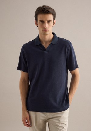 Giovane uomo con capelli corti castani che indossa una polo blu navy a maniche corte e pantaloni beige, in piedi davanti a uno sfondo chiaro e uniforme.