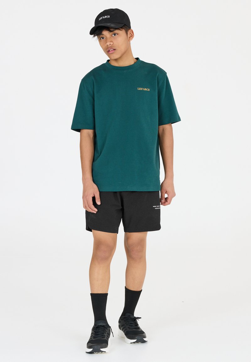 Mörkgrön t-shirt med kort ärm och guldtext, kombinerad med svarta shorts, svart keps, svarta strumpor och svarta sportskor.