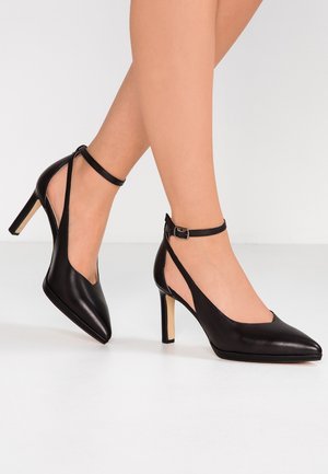 Sorte læder højhælede sko med spidse tæer og udskårne sider. Har en ankelrem med spænde. Elegant og strømlinet design.
