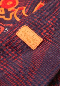 Rood en marineblauw geblokt stof met een oranje rubberen logo patch. De textuur is zacht met een gebreide uitstraling, wat de levendige kleuren benadrukt.