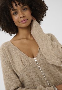 Bežový pletený sveter s hlbokým V-výstrihom, ktorý má textúrované vzory a dekoratívne gombíky pozdĺž prednej strany. Mäkká, útulná tkanina.