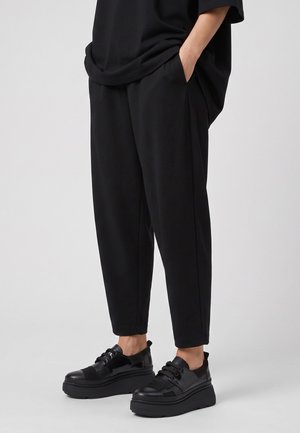 Person trägt lockere schwarze Hosen und schwarze Plateausneaker, steht mit den Händen in den Taschen vor einem schlichten hellen Hintergrund.