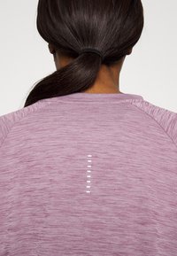 Lila Sportoberteil aus strukturiertem Stoff. Es verfügt über ein einzigartiges Designelement: weiße vertikale Linien, die mittig auf dem Rücken positioniert sind.