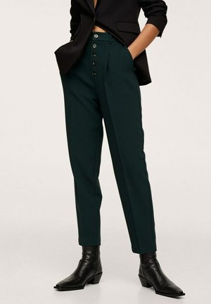 Donkergroen gepolieste broek met een hoge taille, voorzien van een rij zwarte knopen en scherpe plooien. Gecombineerd met zwarte enkelboots.