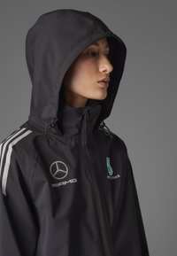 Sort vandtæt jakke med lynlås foran, justerbar hætte, sølvfarvede detaljer og logoer, herunder Mercedes og Petronas, på brystet.