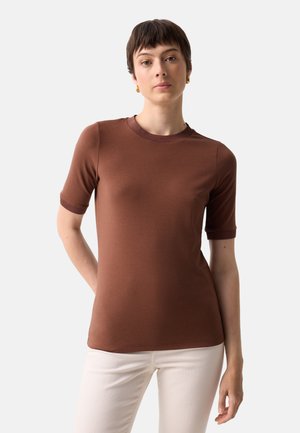 Femme aux cheveux brun courts portant un t-shirt marron ajusté à manches courtes et un pantalon blanc, debout devant un fond clair uni.