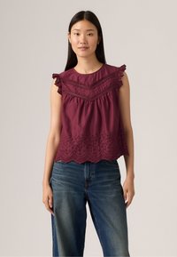Top bordeaux senza maniche con bordino in pizzo, corpetto a trame, e orlo a scallop con ricami floreali, abbinato a jeans in denim blu.
