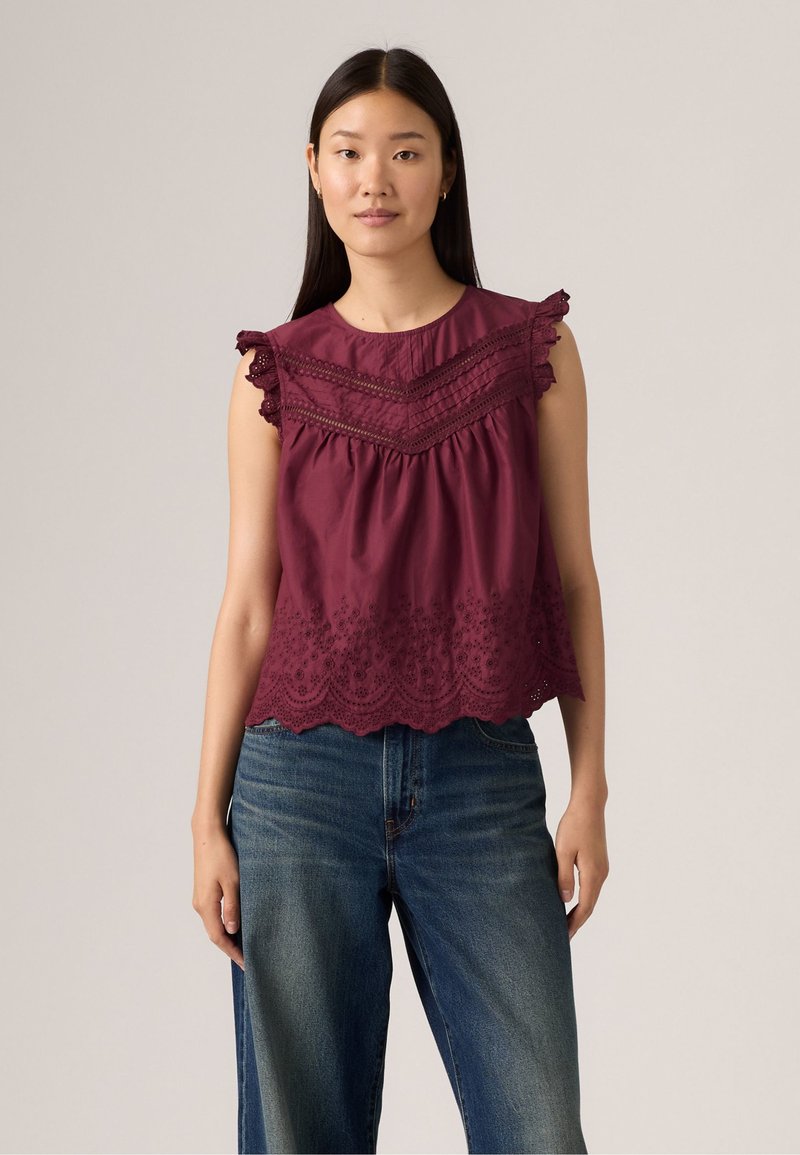 Top bordeaux senza maniche con bordino in pizzo, corpetto a trame, e orlo a scallop con ricami floreali, abbinato a jeans in denim blu.