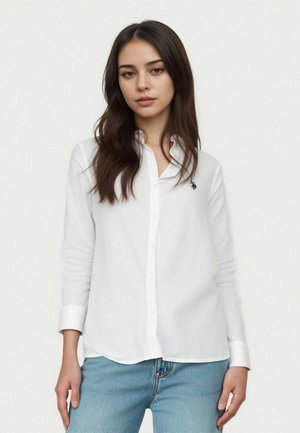 Giovane donna con lunghi capelli castani che indossa una camicia bianca con bottoni e jeans blu, su uno sfondo chiaro e semplice.