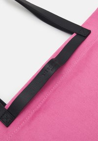 Różowa torba płócienna na ramię z czarnymi skórzanymi uchwytami, o teksturowanym wykończeniu i subtelnym, wytłoczonym logo marki na uchwycie.