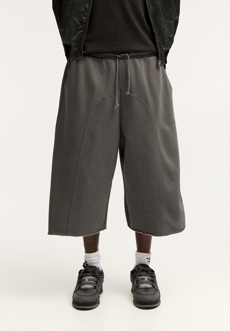 Grå överdimensionerade shorts i mjukt tyg, med elastisk midja, dragsko och svepande sömdetaljer. Bärs med svarta sneakers.
