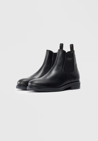 Une paire de élégantes bottes Chelsea en cuir noir avec des panneaux élastiques sur les côtés et des languettes de tirage, marquées "GANT" sur la cheville extérieure.