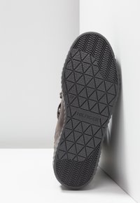 Semelle d'une basket gris foncé avec un motif géométrique et le logo "HILFIGER", placée contre un mur d'angle blanc.
